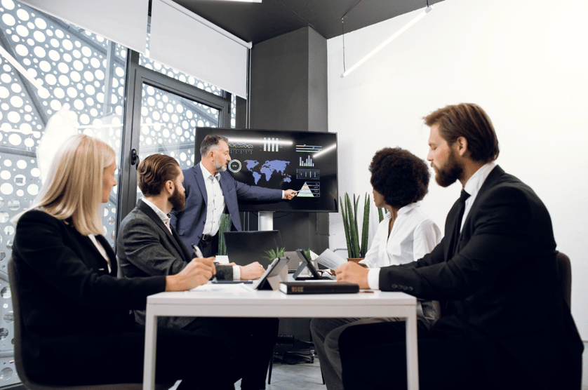 La imagen muestra un grupo de inversionistas expertos financieros multiétnicos, sentados a la mesa en la oficina, mientras emprendedor de mediana edad les presenta gráficos y mapas sobre la idea de negocio y la actividad de la empresa en el televisor de pared.