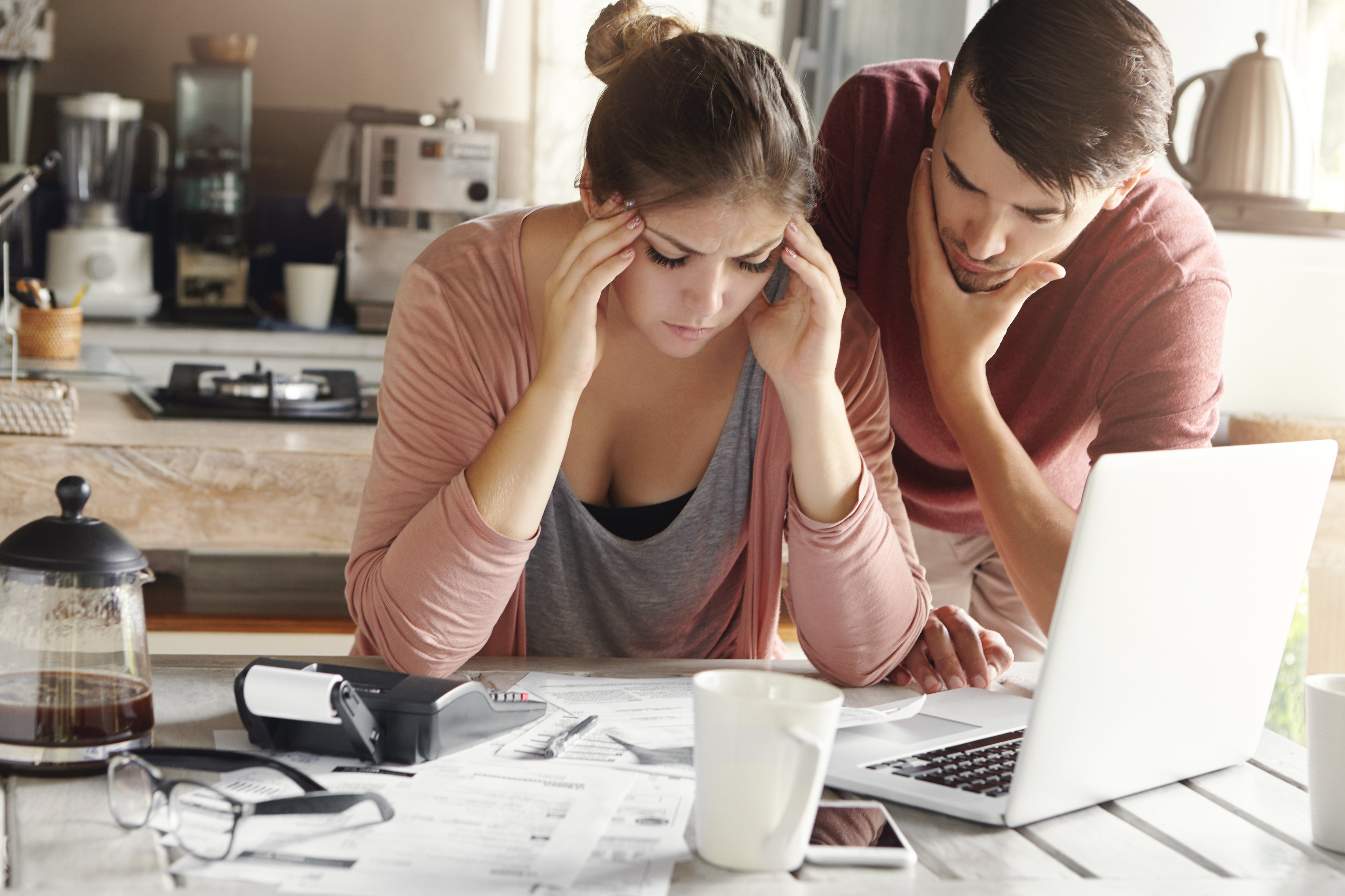 Mujer joven con expresión de desánimo y dolor de cabeza, sosteniendo sus sienes con las manos. Su rostro refleja el estrés y la preocupación causados por la falta de dinero para pagar las deudas familiares, crédito malo, deuda de tarjetas. A su lado, su marido muestra desconcierto mientras busca una solución. La imagen captura la angustia y la presión financiera que enfrenta la pareja, poca capacidad de pago resaltando la importancia de abordar y buscar alternativas para superar las dificultades económicas.